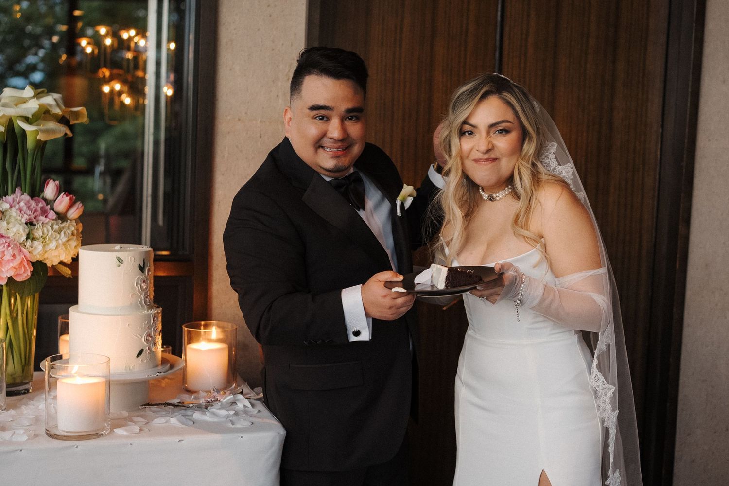A newlywed couple shares their first bite of wedding cake together at their reception beside glowing candles and flowers.