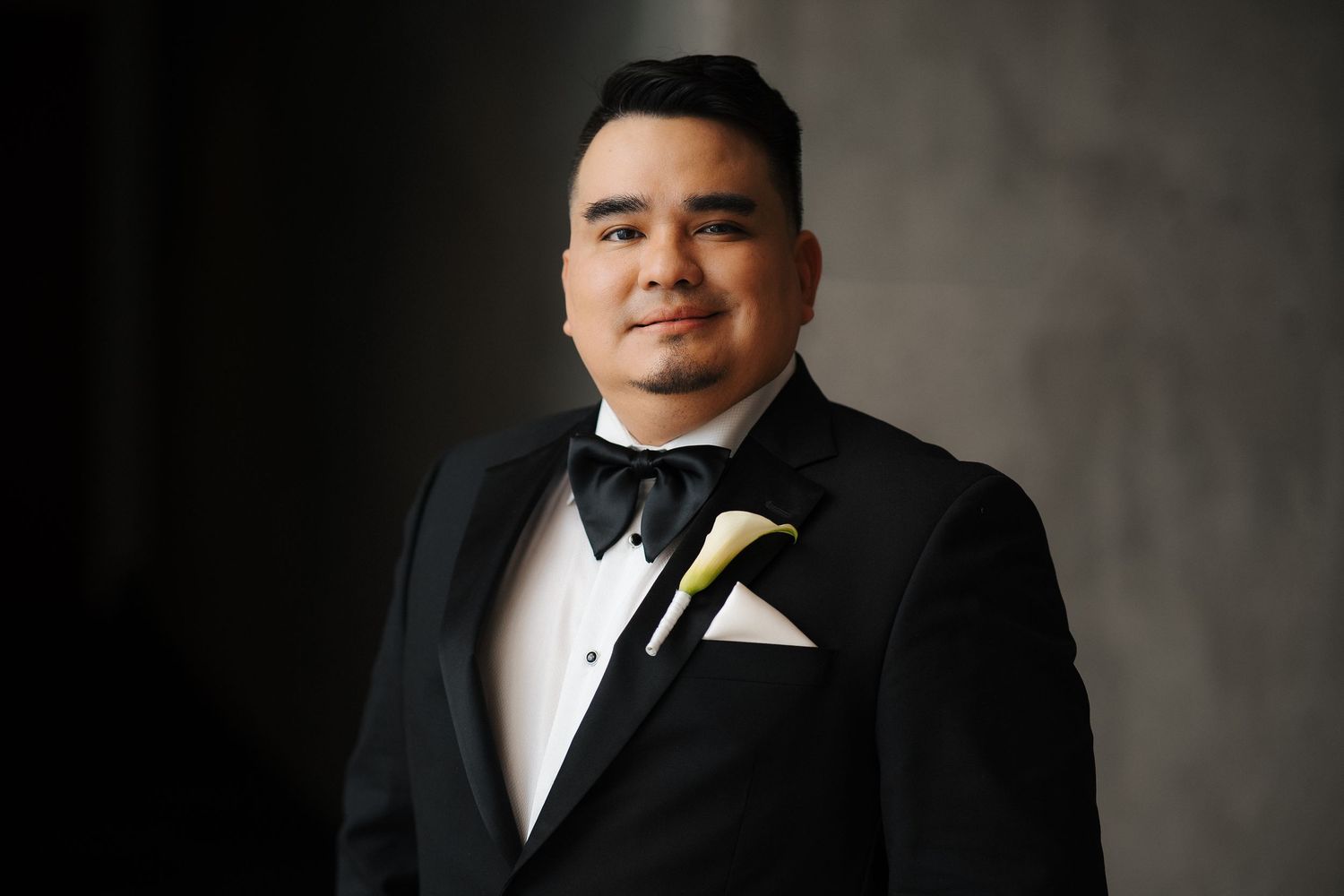 Portrait of someone in a formal black tuxedo with bow tie and white calla lily boutonniere against dark background.