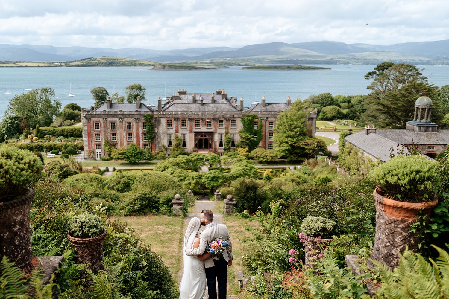 A couple embraces in a garden overlooking a grand historic mansion and scenic coastal bay in Ireland.
