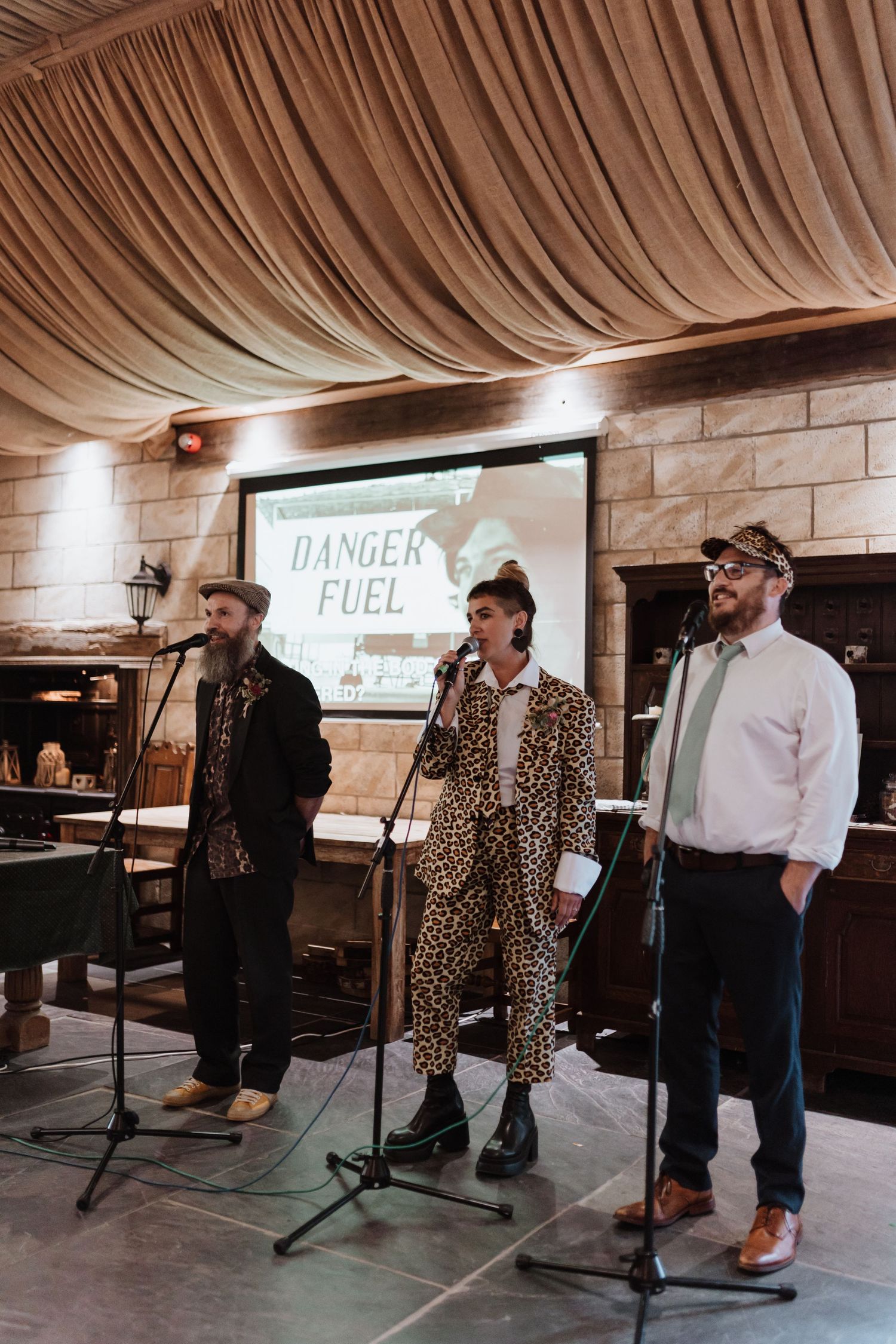 Musicians perform on stage in a rustic venue with wooden walls and minimal lighting while wearing casual attire.