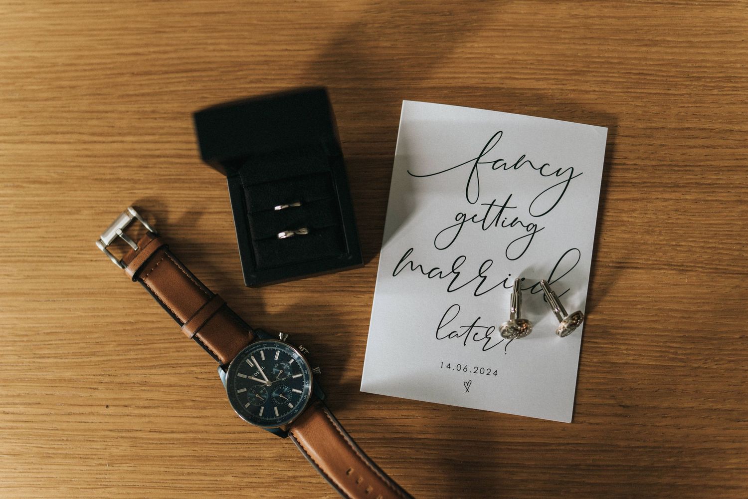 Brown leather watch with black face lies next to wedding ring box and calligraphy card on wooden surface.