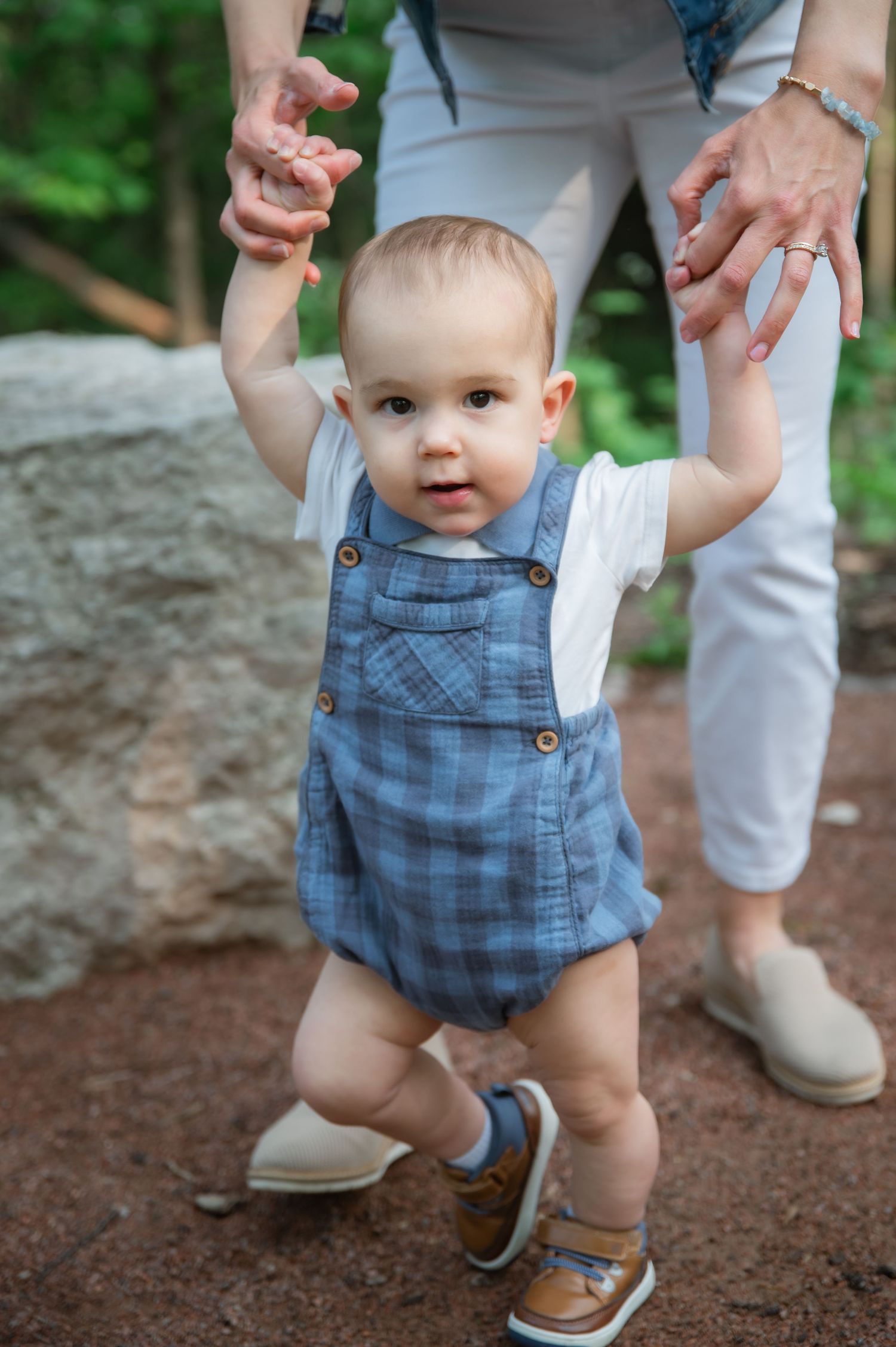 Small child in blue plaid overalls takes first steps while being supported at the wrists on a dirt path.