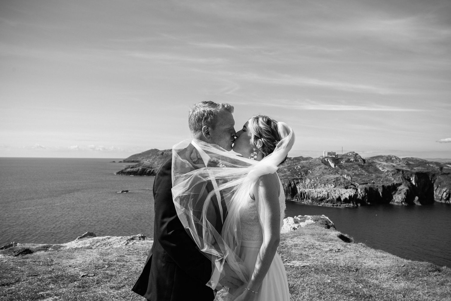 A veil floats dramatically in the wind on a scenic coastal cliff overlooking the ocean in black and white.
