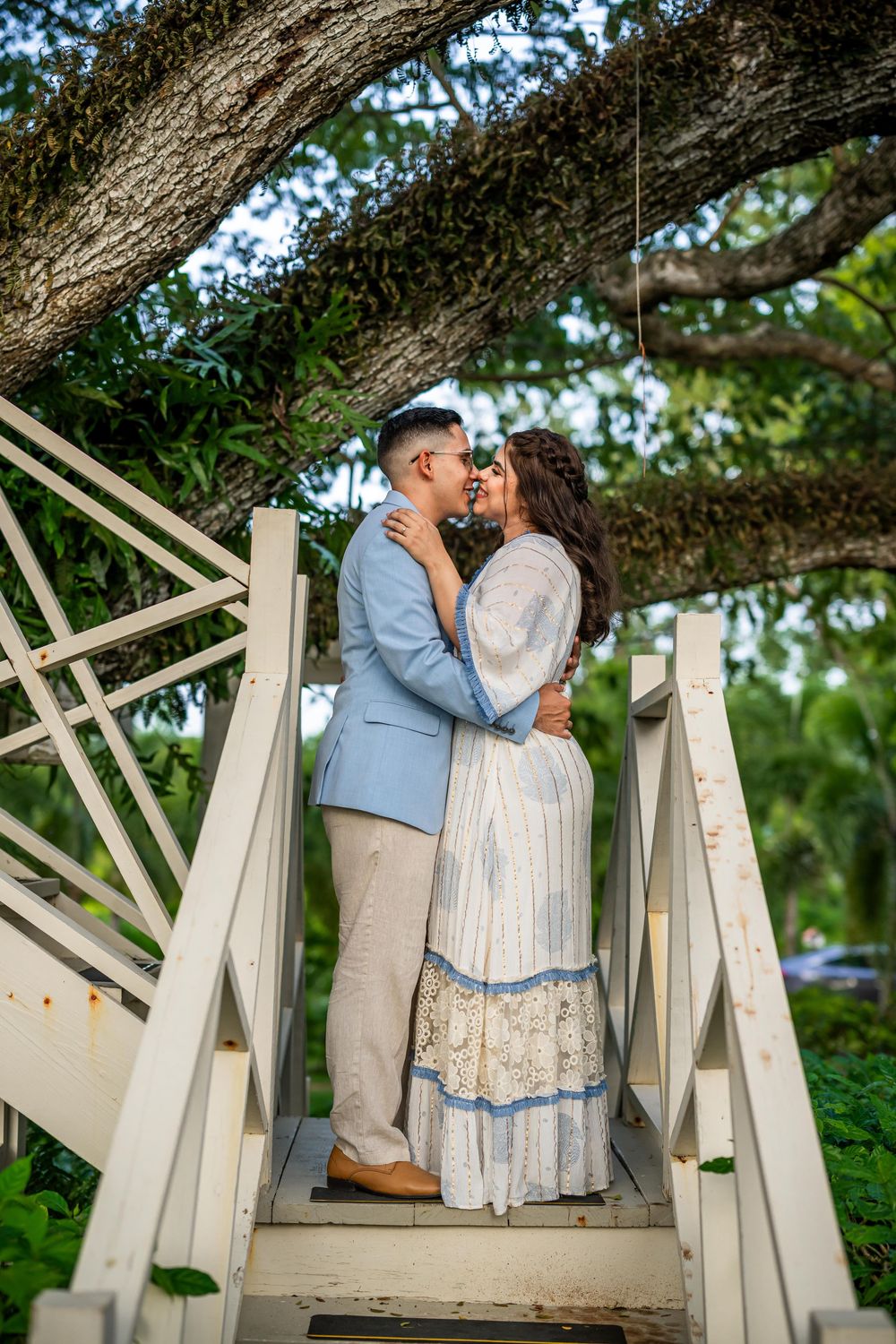 Romantic couple in vintage formal attire embrace on white wooden bridge steps under a large tree.