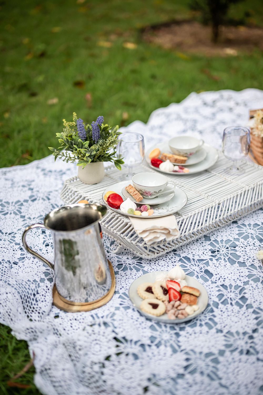 Vintage outdoor tea party setting with lace tablecloth, silver pitcher, and fresh pastries on a rustic wooden board.