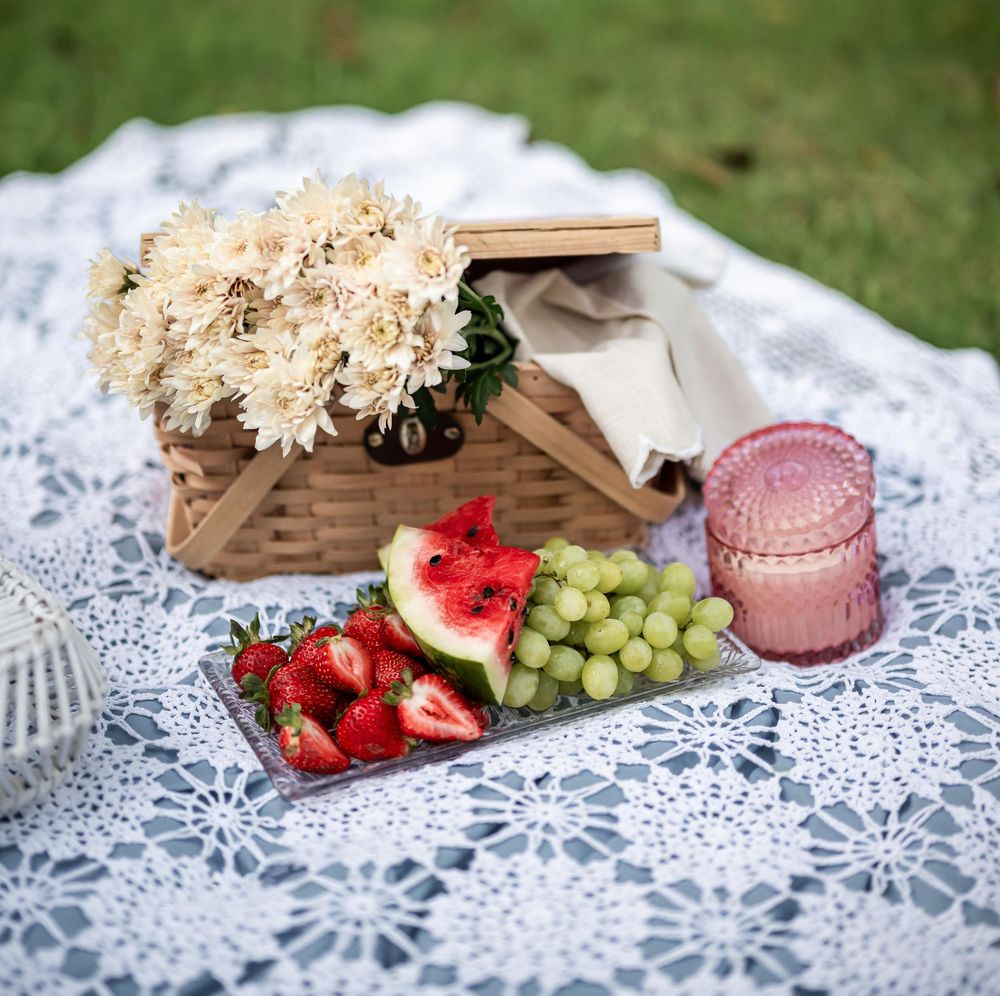 A picnic setup with white daisies, fresh fruit, and pink drinks on a delicate lace tablecloth outdoors.