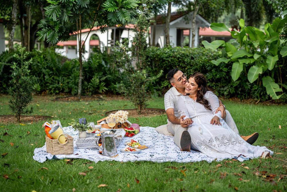 Romantic outdoor picnic setup on white blanket with flowers and food spread in lush garden setting.
