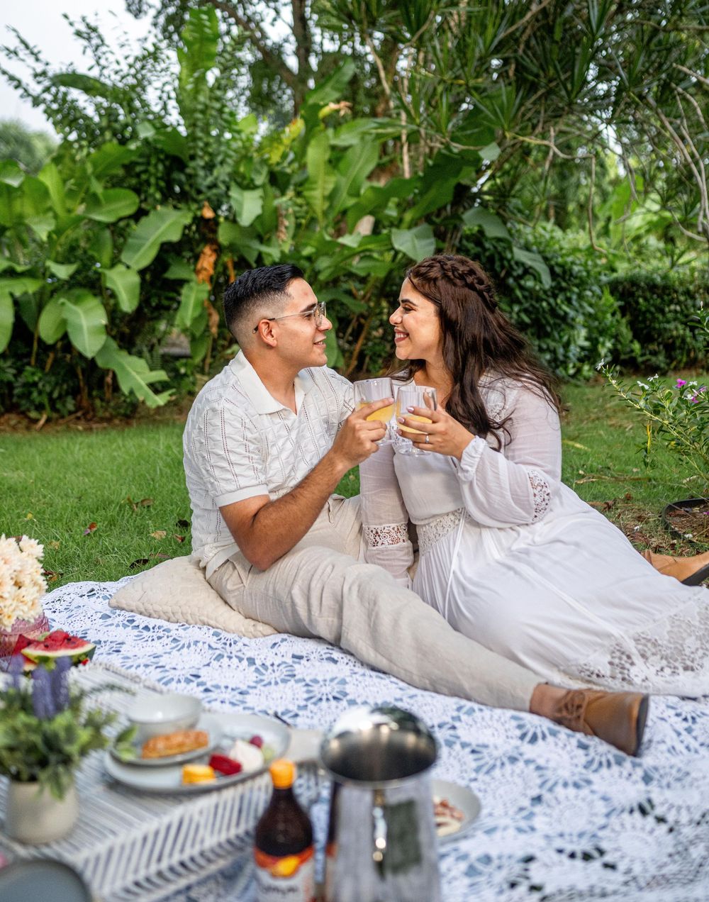 Couple enjoying a romantic outdoor picnic on a white lace blanket with wine and snacks in a lush garden setting.