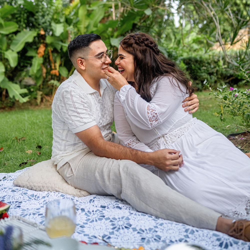 A romantic couple enjoys a picnic date while sitting on a white lace blanket in a garden setting.