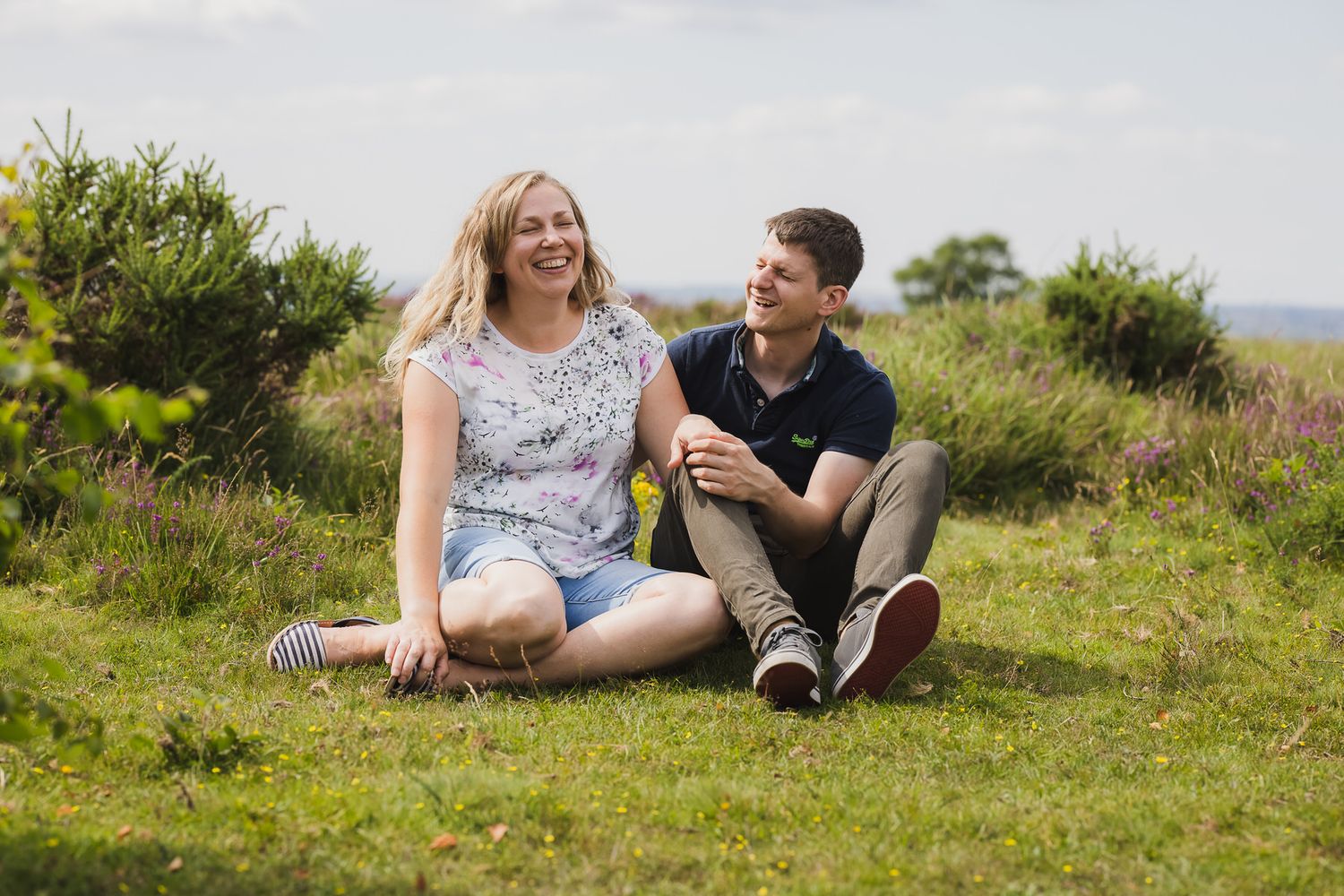 Two people sit together on grassy hill sharing joyful moment in nature on sunny day.