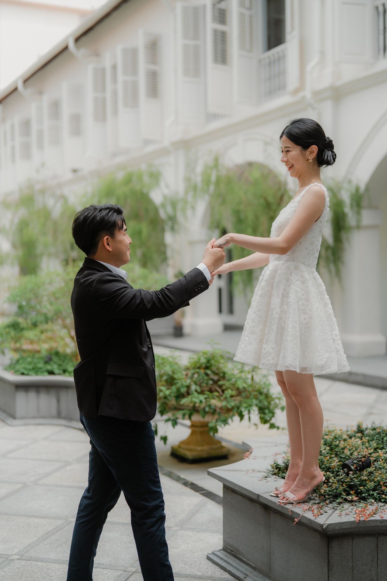 A couple in wedding attire playfully poses in a white courtyard garden with elegant architectural arches.