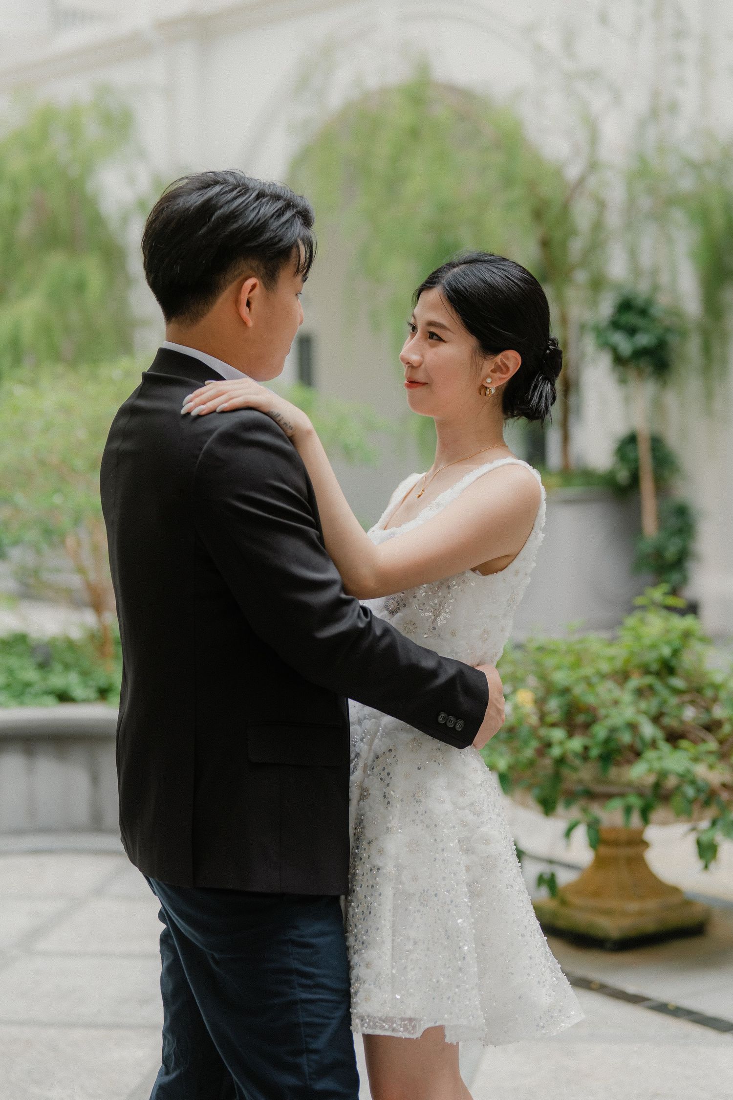 Couple in wedding attire embracing in courtyard with white architecture and greenery in background.