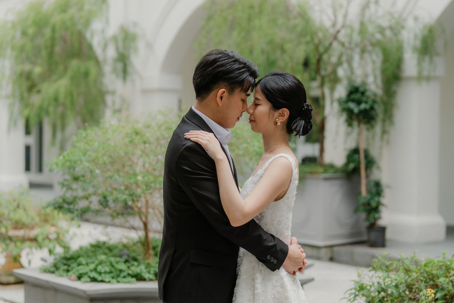 A tender kiss shared between a couple in wedding attire against an elegant white archway and greenery.