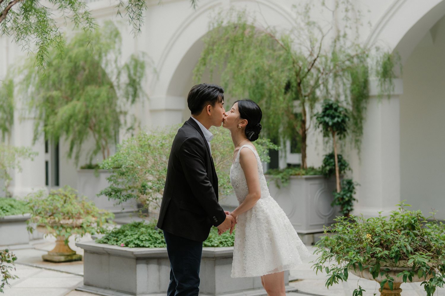 A wedding couple embraces tenderly in a beautiful courtyard setting with white arches and willow trees.
