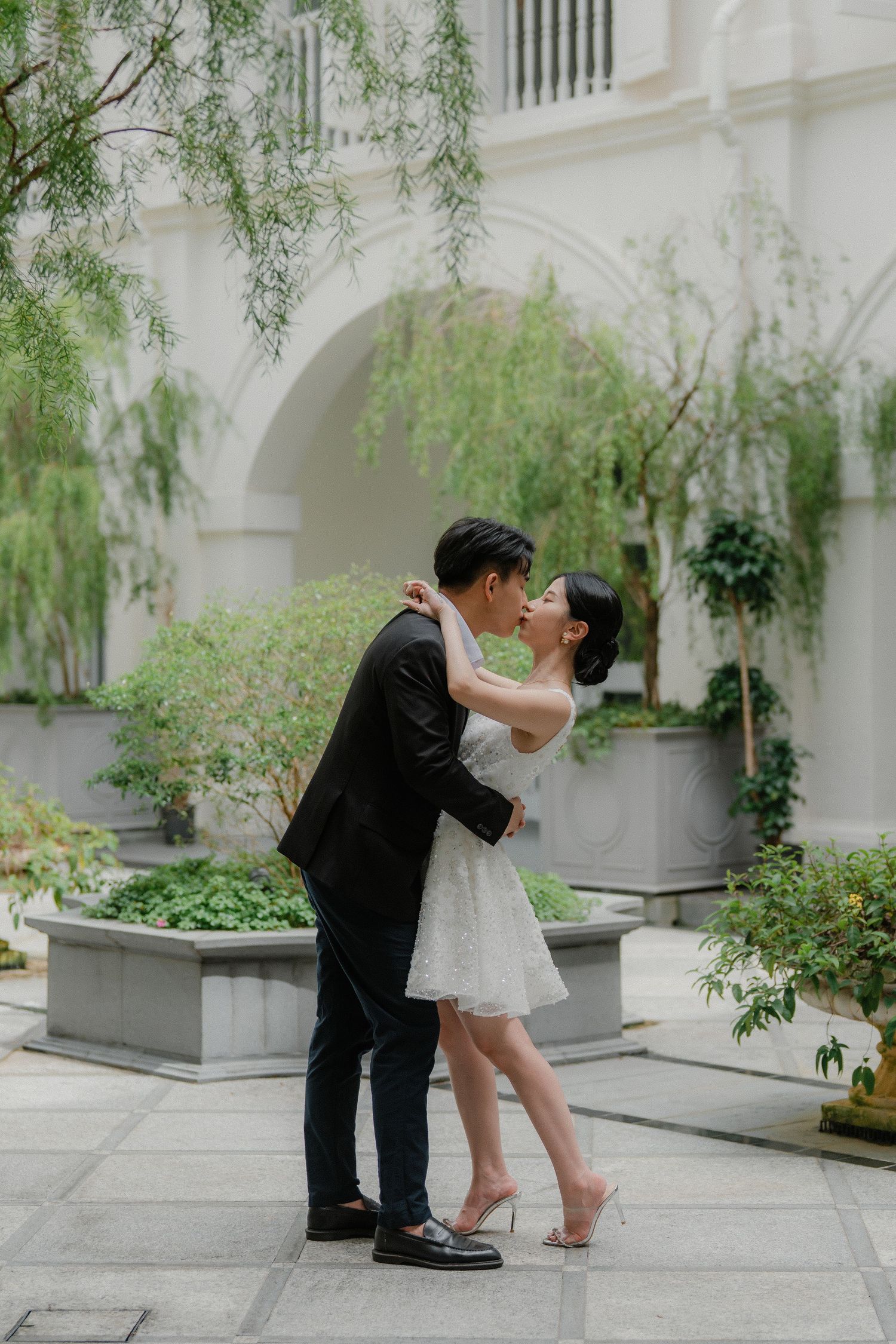 Newlyweds share an intimate moment under a white archway surrounded by willow trees and planters.