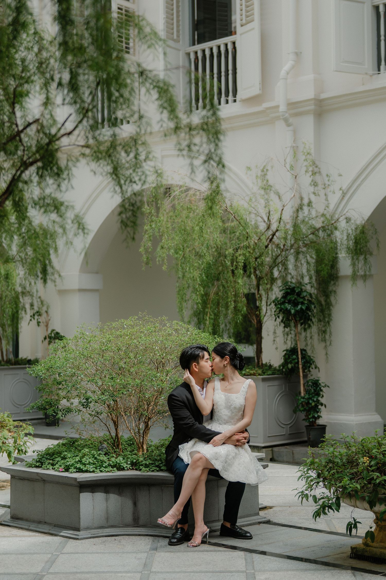 A romantic couple in wedding attire sitting together on a stone bench surrounded by hanging willow trees and arches.