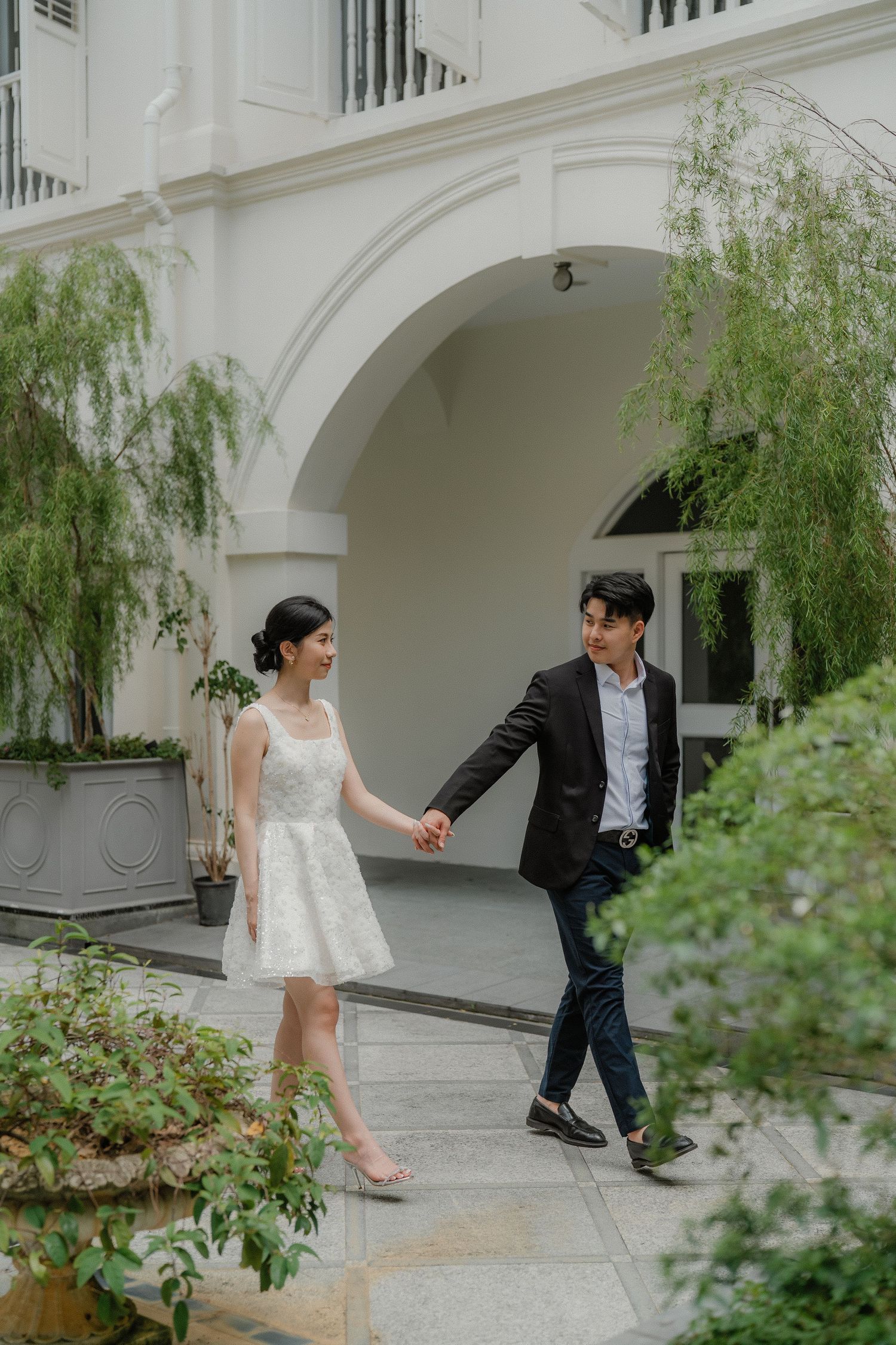 A couple in formal wear walking hand in hand through an elegant white archway surrounded by lush greenery.