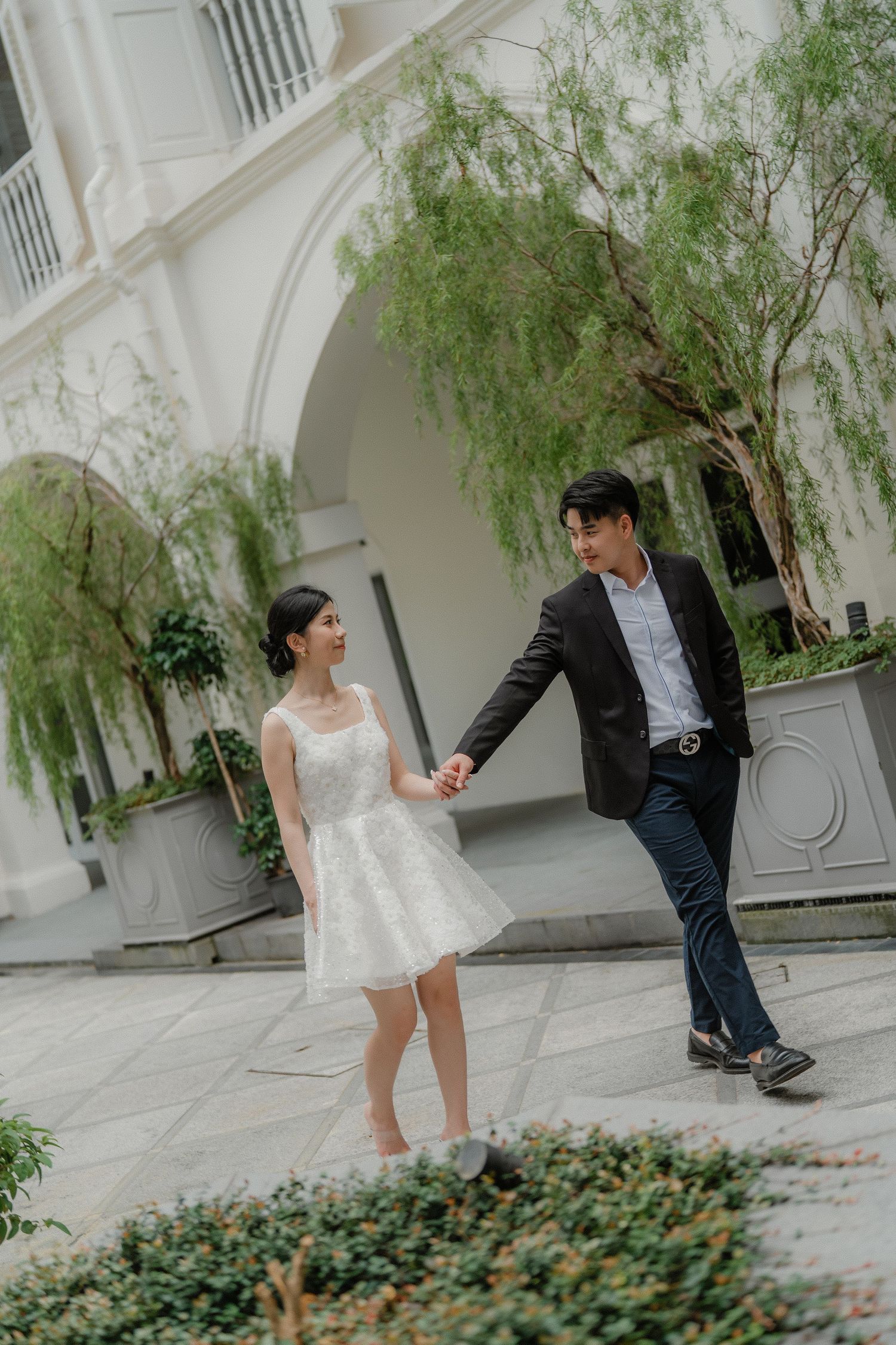 A couple playfully interacts in an elegant white courtyard garden with weeping willows and garden beds.