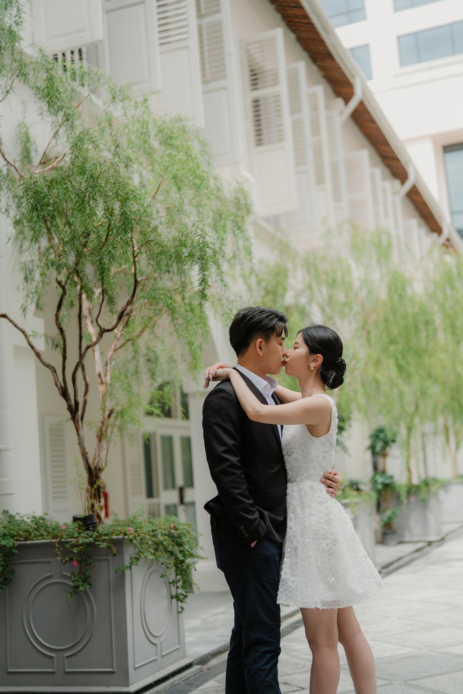 A romantic moment captured between a couple in formal attire embracing in front of a white colonial building with willow trees.