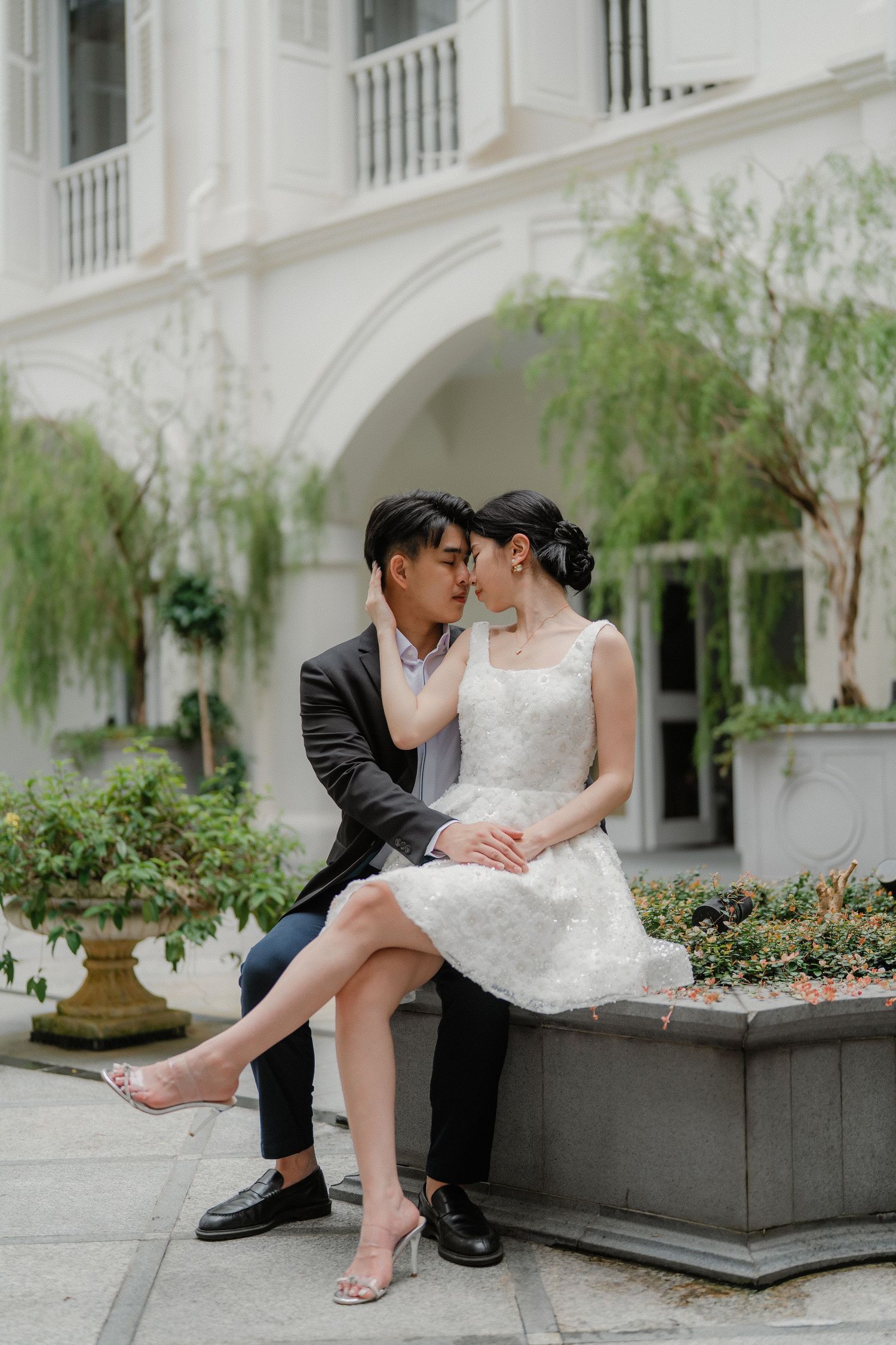 Couple in elegant attire shares an intimate moment on stone bench in front of white arched building.