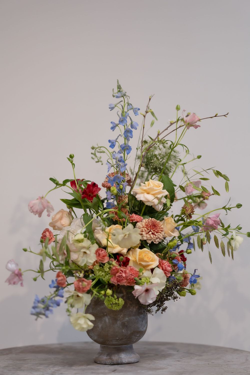 Lush floral arrangement in a stone urn with pastel roses, blue delphiniums, pink carnations, and trailing greenery.