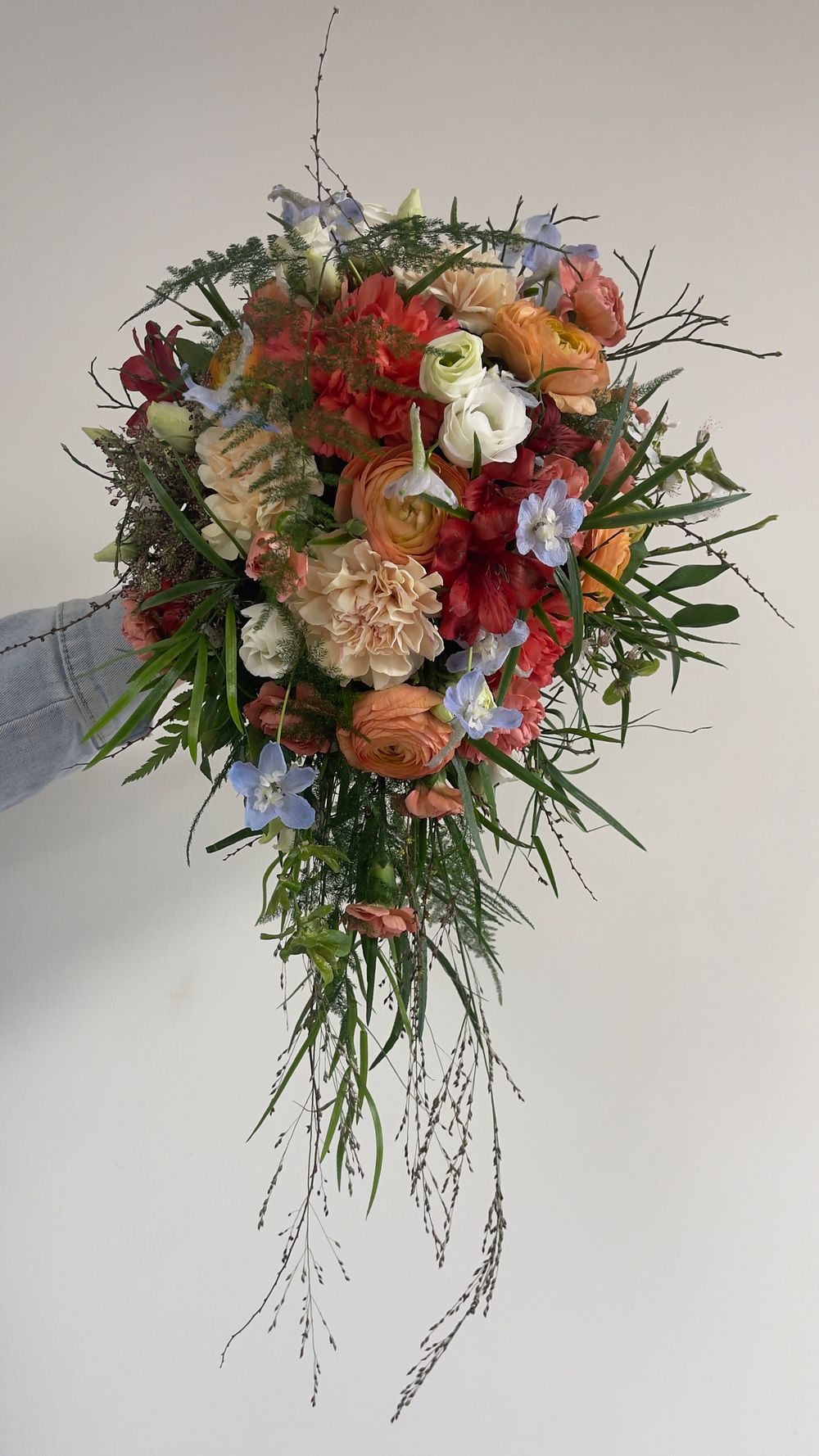 Hand-held cascading bridal bouquet with orange ranunculus, red flowers, blue delphinium, and trailing greenery.