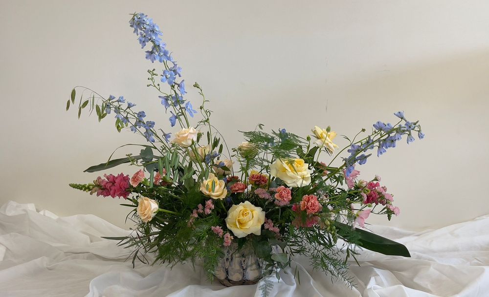 Low garden-style floral centerpiece with yellow roses, blue delphiniums, pink alstroemeria, and wild greenery.