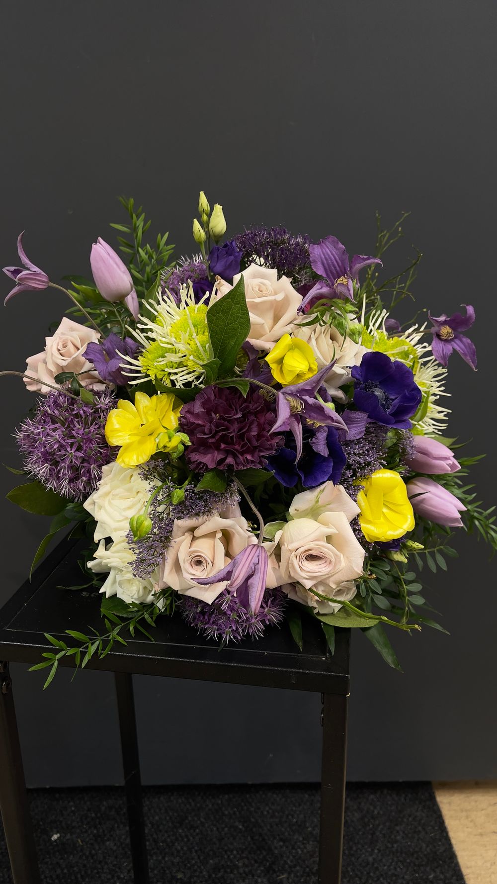 Vibrant purple and yellow bouquet with roses, anemones, allium, tulips, and lush greenery on a stand.