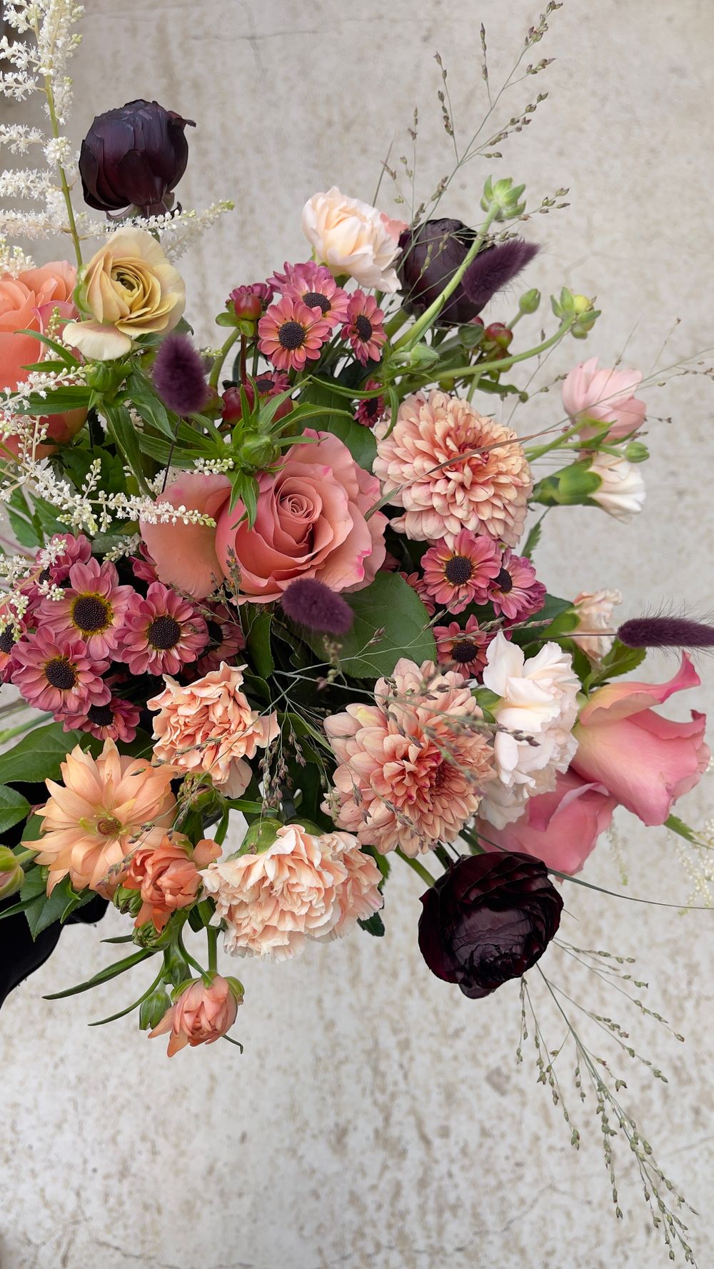 Rich bouquet of blush roses, dark burgundy tulips, peach dahlias, pink daisies, and wispy grasses.
