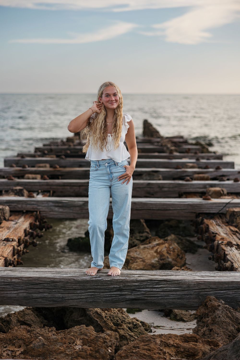 A person in casual summer attire stands barefoot on wooden beams by the ocean at sunset.