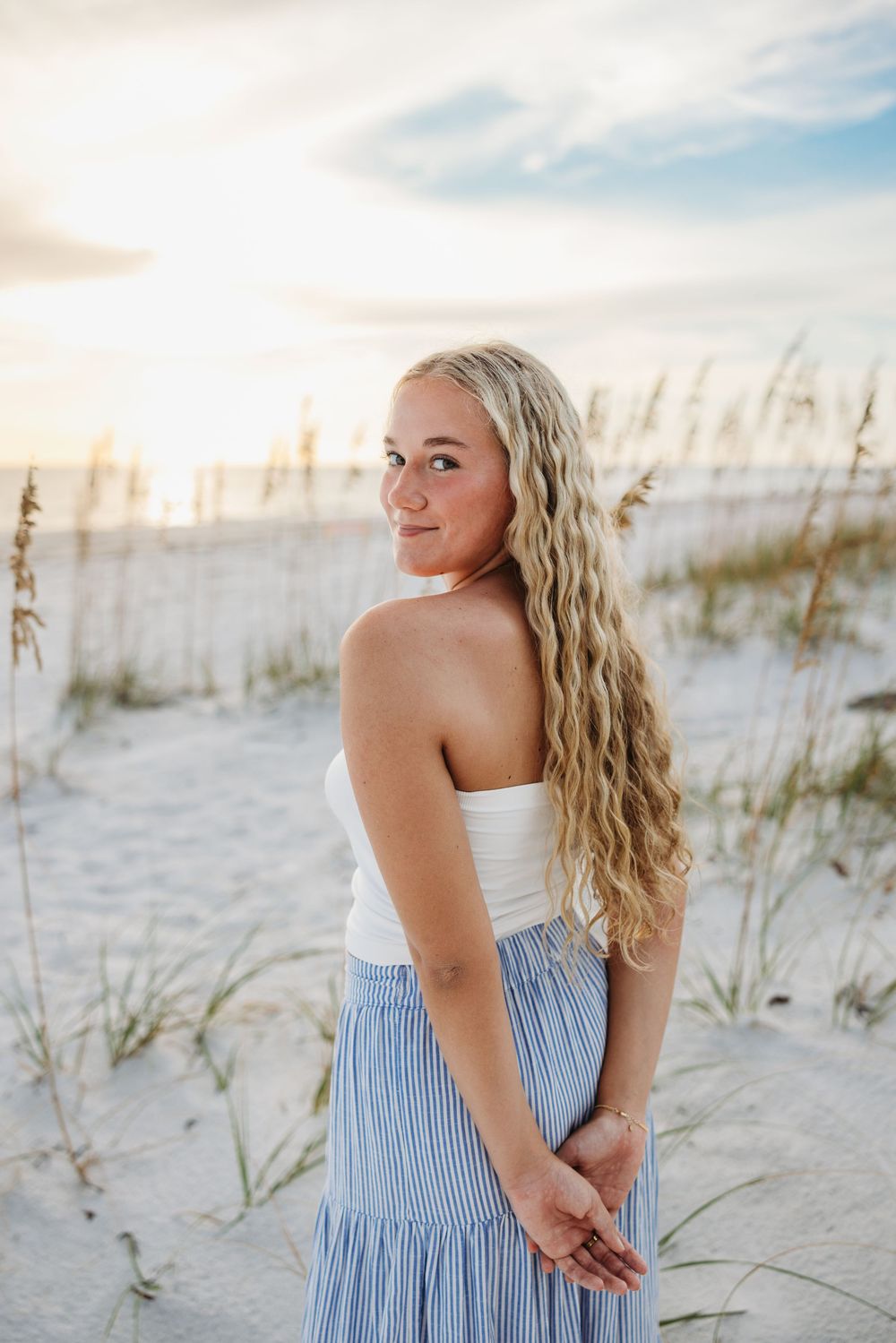 A person in a white top and striped skirt poses on a sandy beach with beach grass and dunes during sunset.