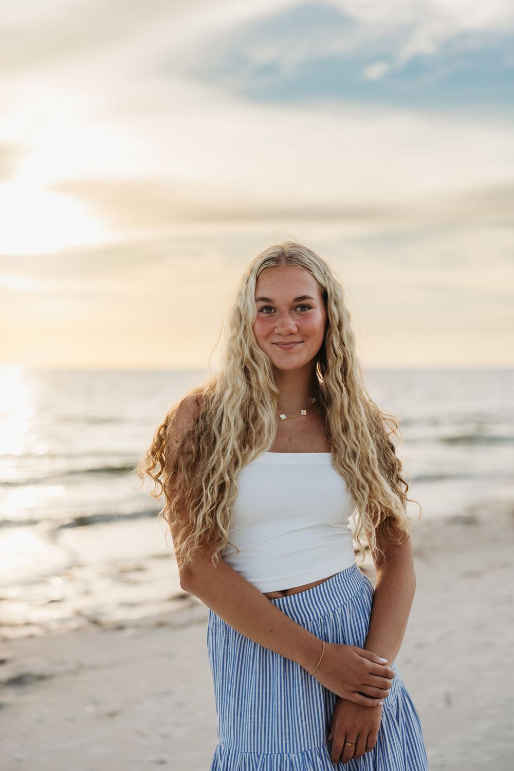 Young model in white crop top and blue striped skirt poses on beach at sunset with waves and cloudy sky in background.