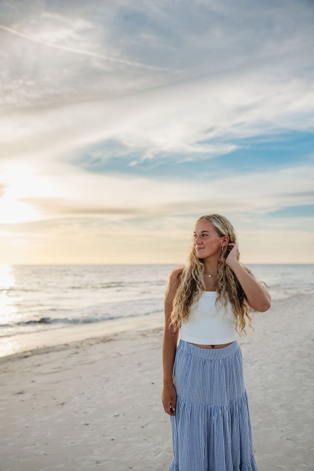 Person in white top and blue flowy skirt stands on beach at sunset with long hair blowing in breeze.