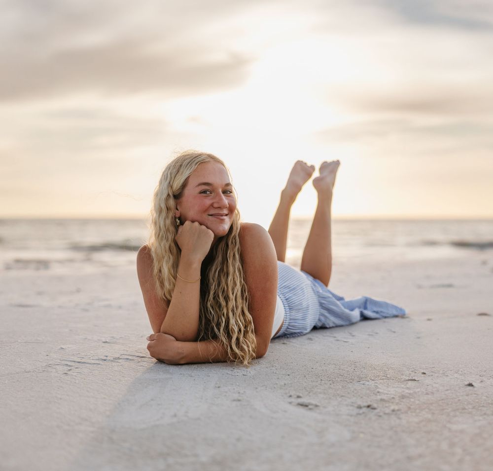 A person relaxing on a sandy beach at sunset while lying on their stomach with feet crossed in the air.