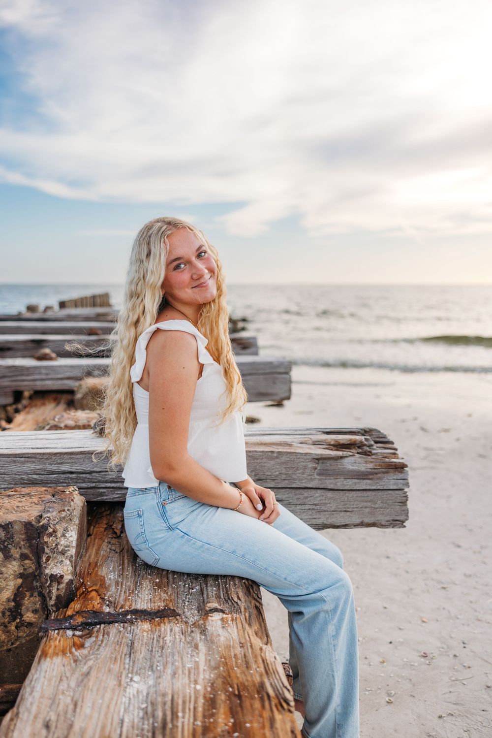 Person wearing white tank top and jeans sits on weathered wooden beams at beach during sunset.