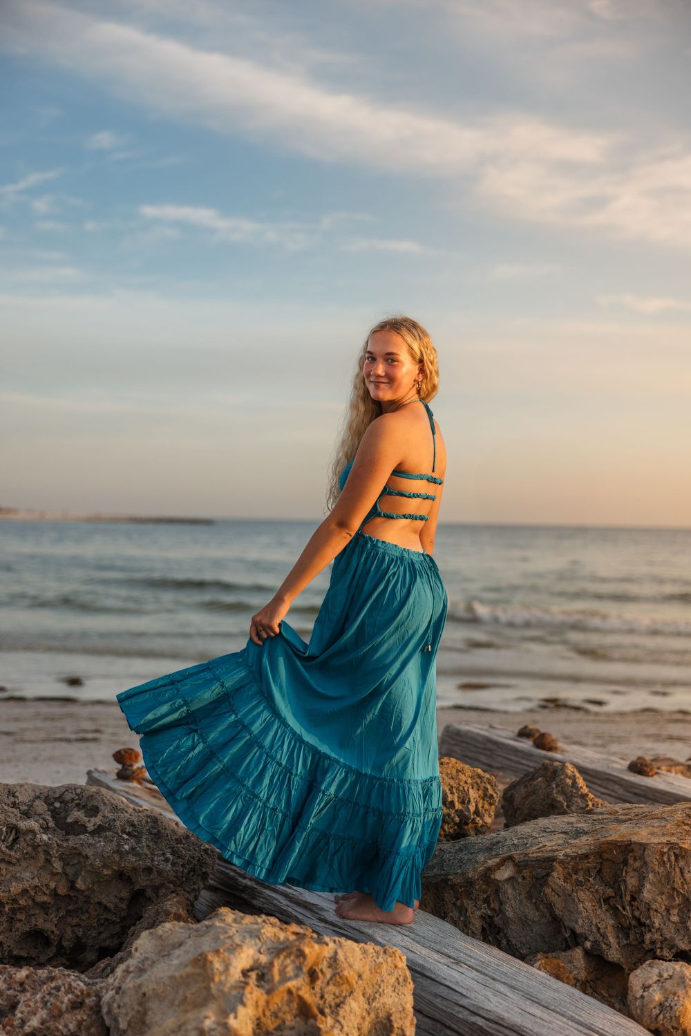Woman in teal dress twirling on rocky beach at sunset with ocean waves in background.