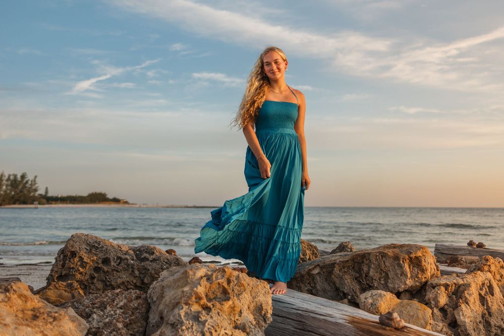 A flowing teal strapless dress catches the ocean breeze as someone poses on rocky coastal shoreline at sunset.
