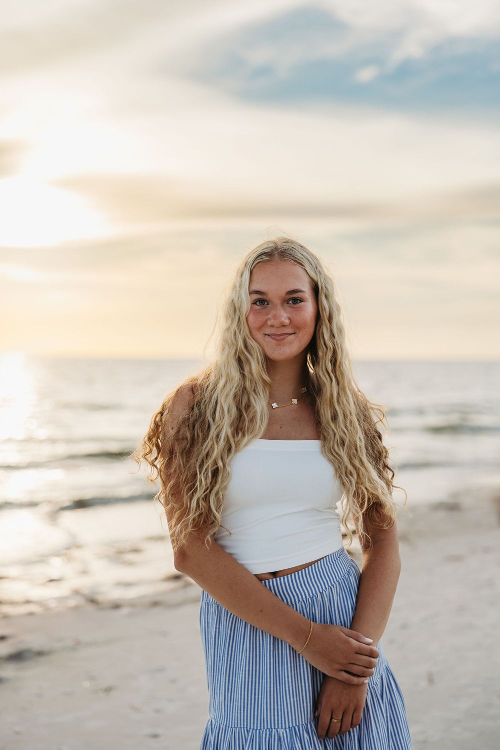 A person in a white crop top and striped blue skirt poses on a beach at sunset.
