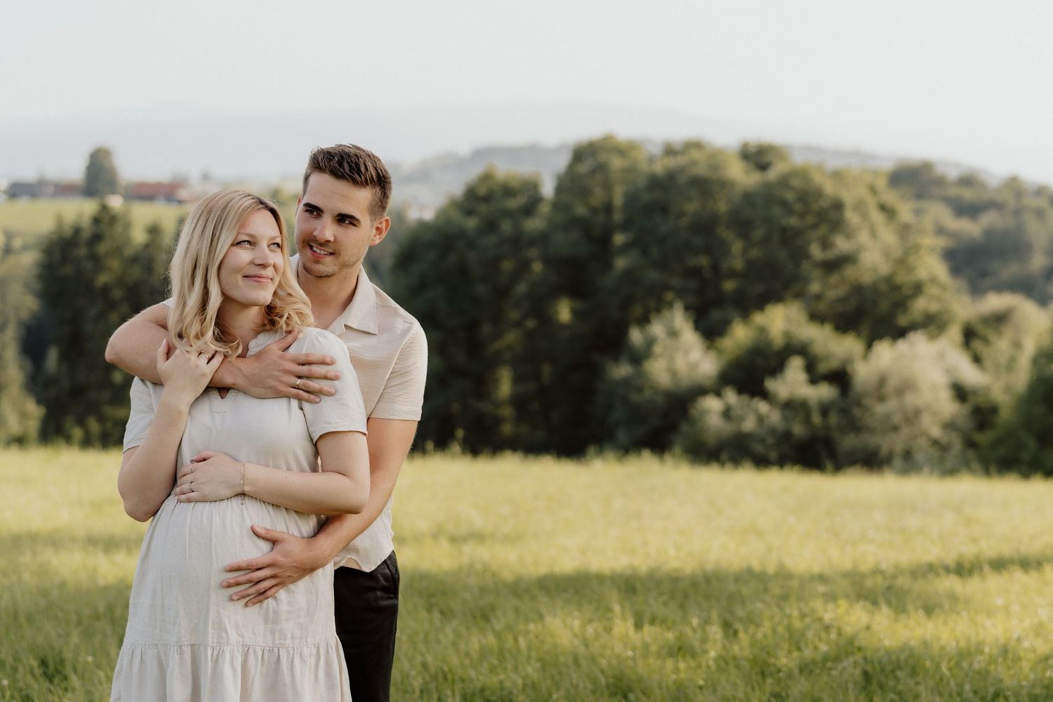 Paar steht Arm in Arm in einer Wiese, beide halten liebevoll den Babybauch