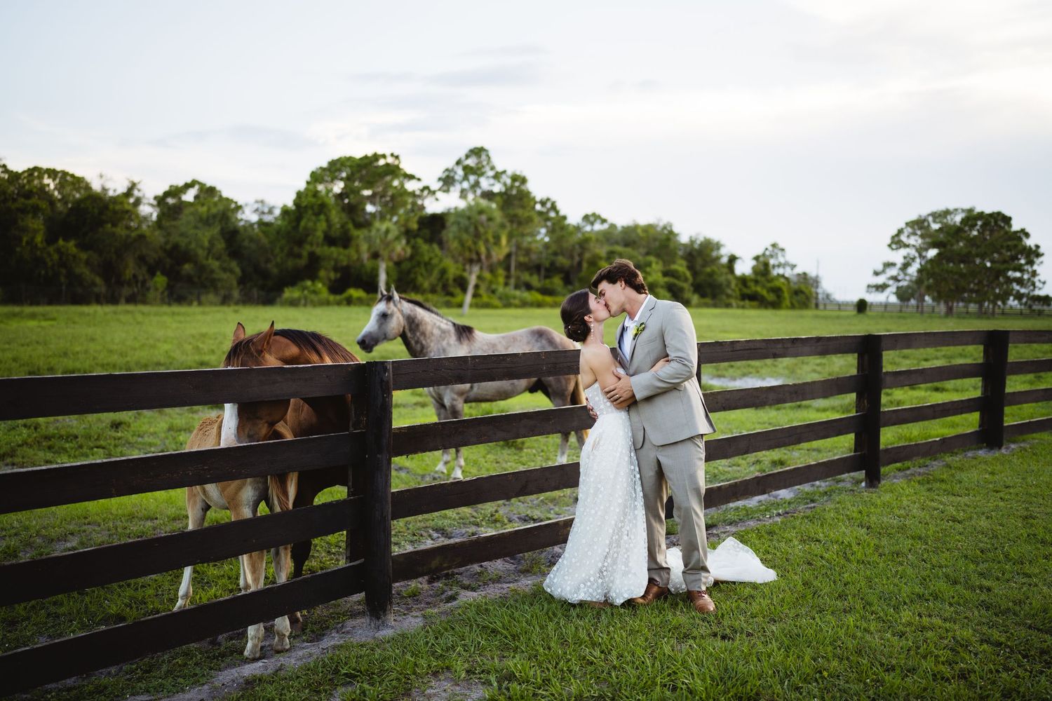 Couple shares intimate moments near rustic wooden fence on green grass at sunset during outdoor wedding photo session.