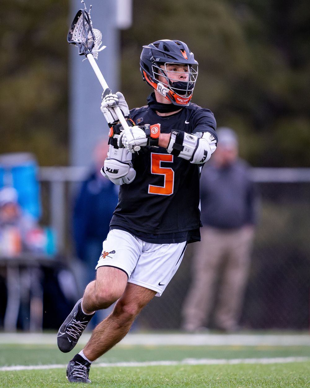 Lacrosse player in black jersey number 5 with protective helmet runs with stick raised during competitive game.