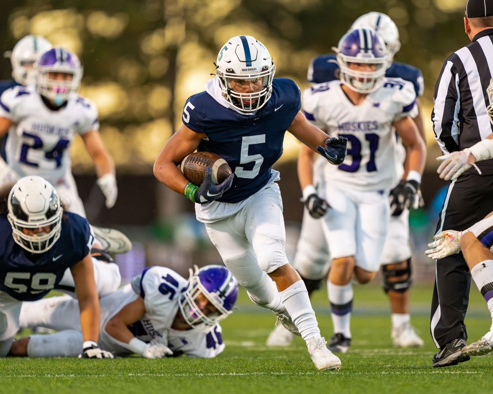 Football player in navy jersey number 5 breaks through the defense during an intense game against white and blue opponents.