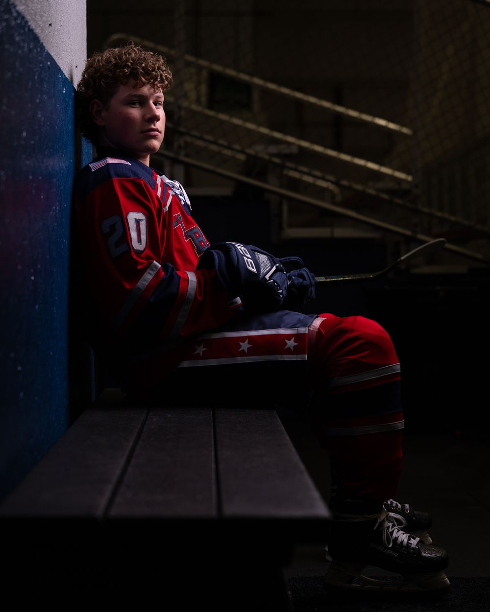 A hockey player in a red and blue uniform sits thoughtfully on a bench in a dimly lit arena.