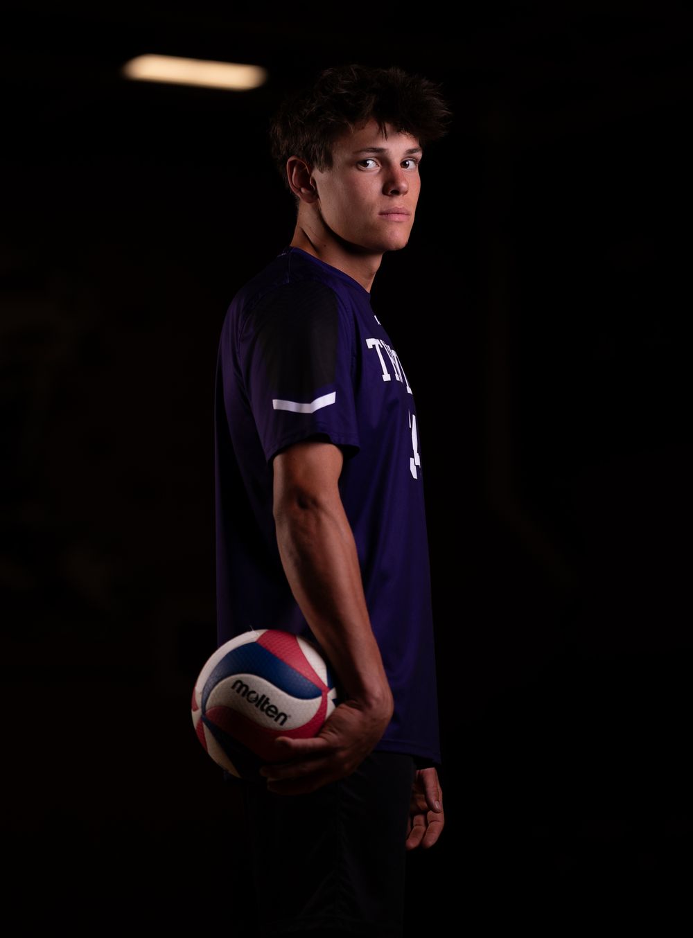 An athlete in a dark navy uniform poses for a dramatic portrait while holding a volleyball against a black background.