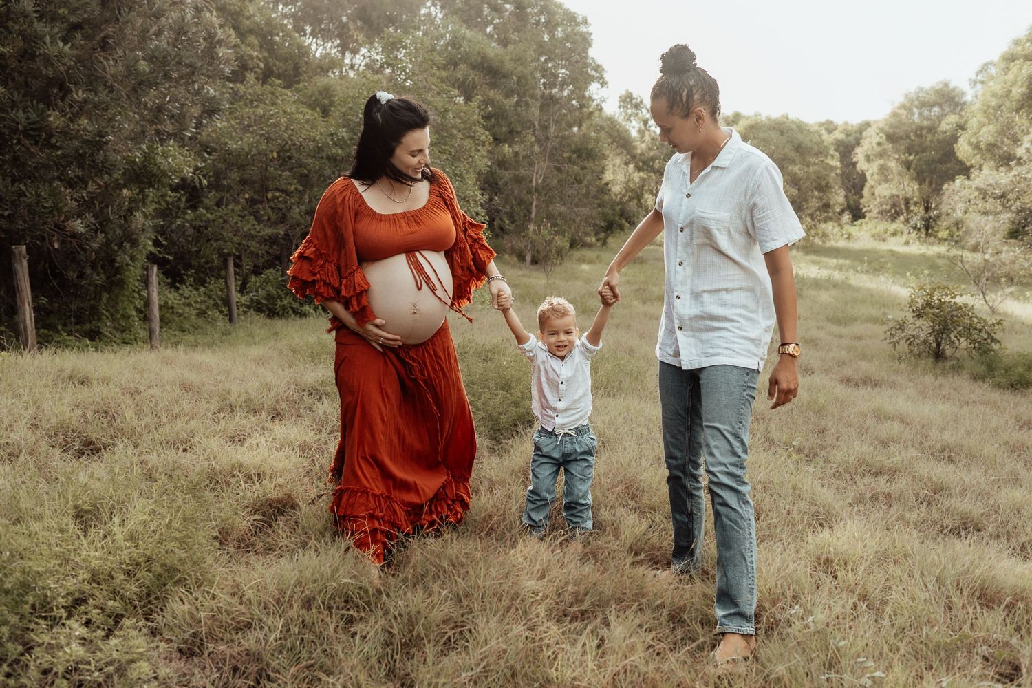 A family walks together through a grassy field, with an expectant mother in a flowing red dress.
