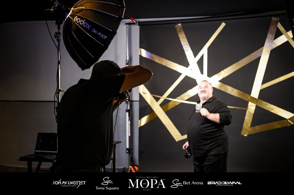 Photographer with studio lighting equipment captures a man posing in front of a gold geometric backdrop at MOPA event.