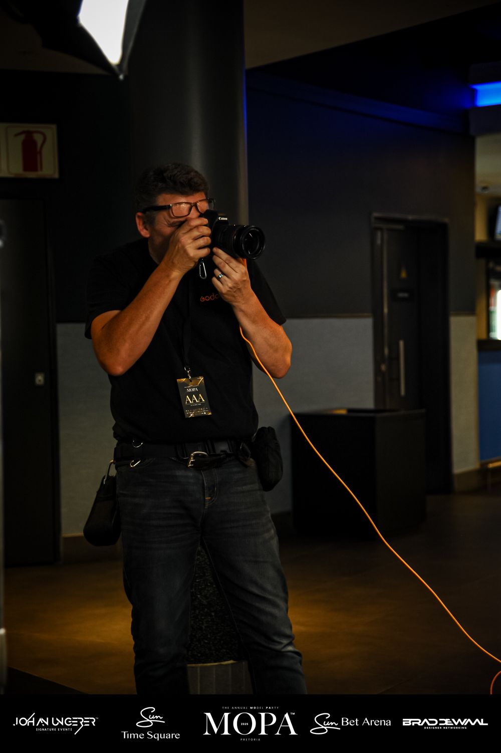 Photographer in black outfit holding camera with red cable strap at MOPA event venue.