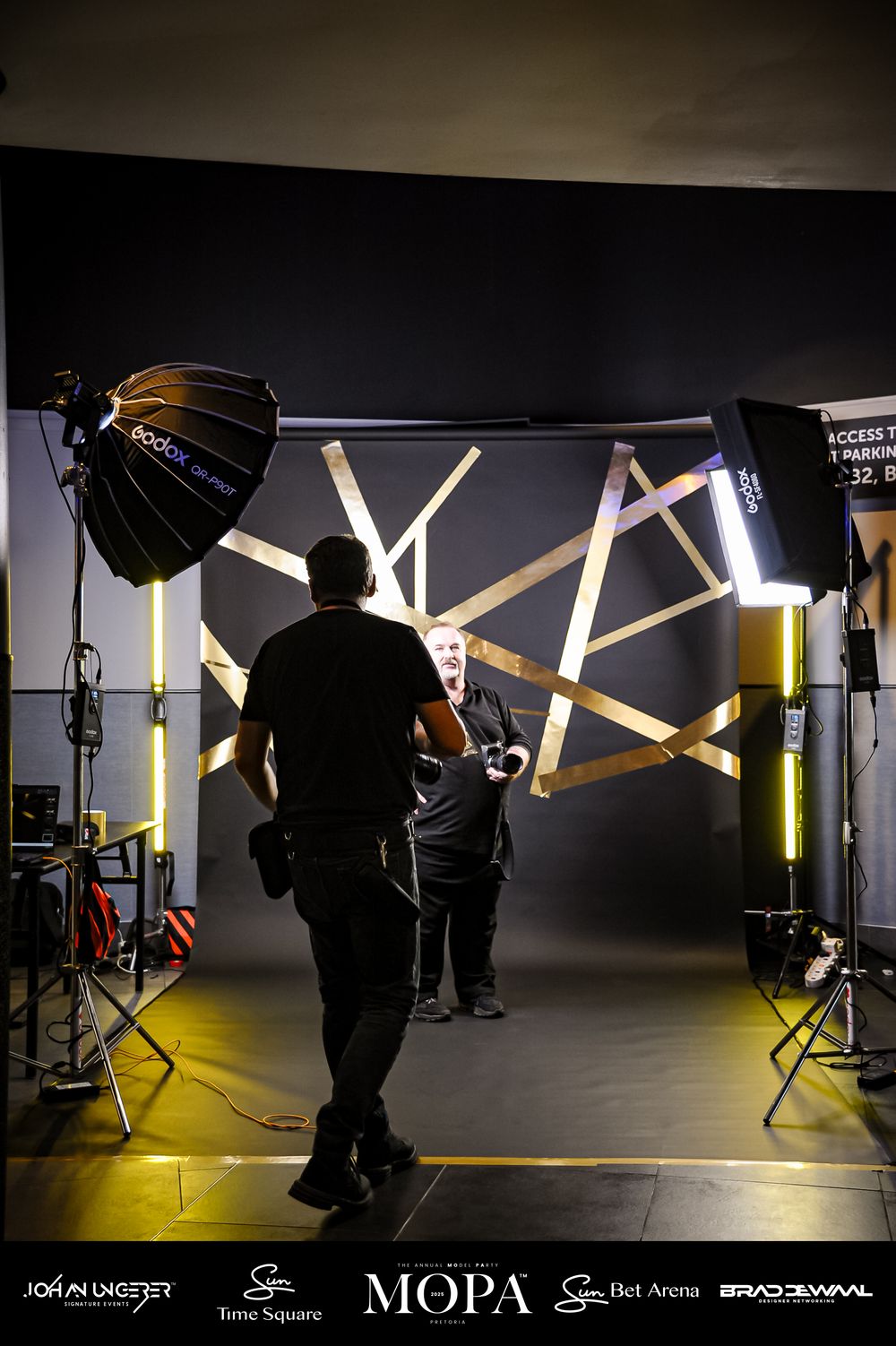Two photographers work in a professional studio with gold geometric backdrop, softbox lighting, and branded sponsor logos.