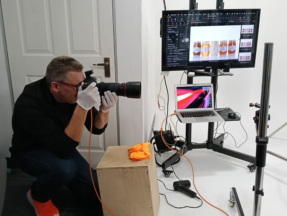 Photographer crouching with DSLR camera in studio setup with dual monitors displaying product photos on adjustable stand.