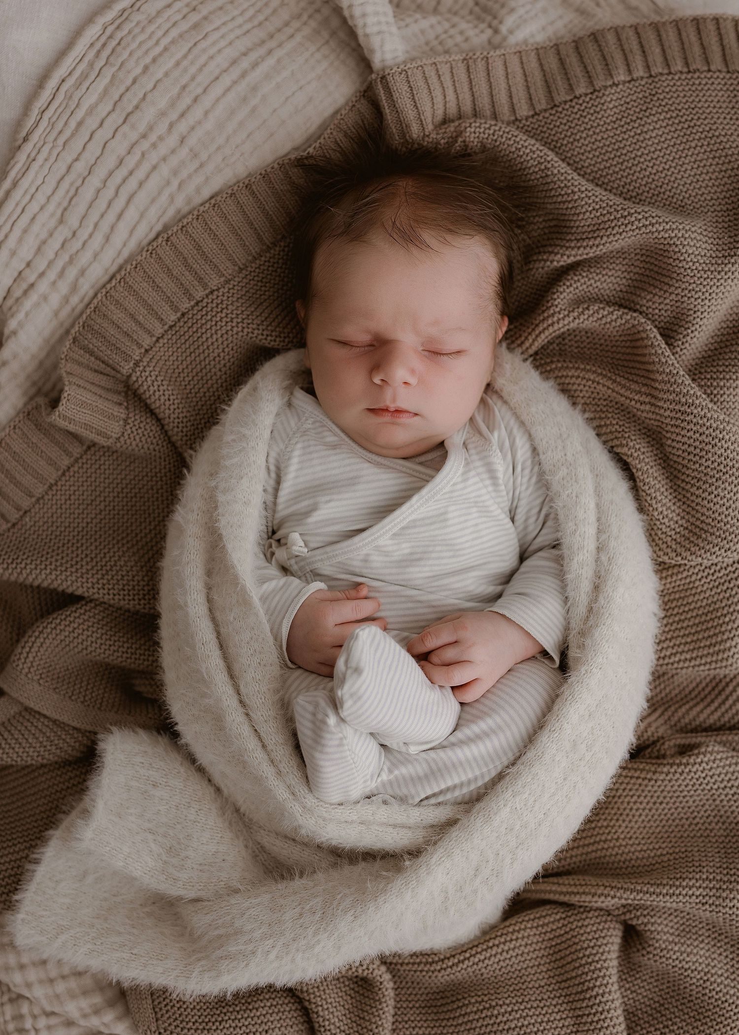 A baby lies swaddled in cozy white blankets against a neutral-toned knit backdrop. Photographed in a studio located in Papamoa, Tauranga